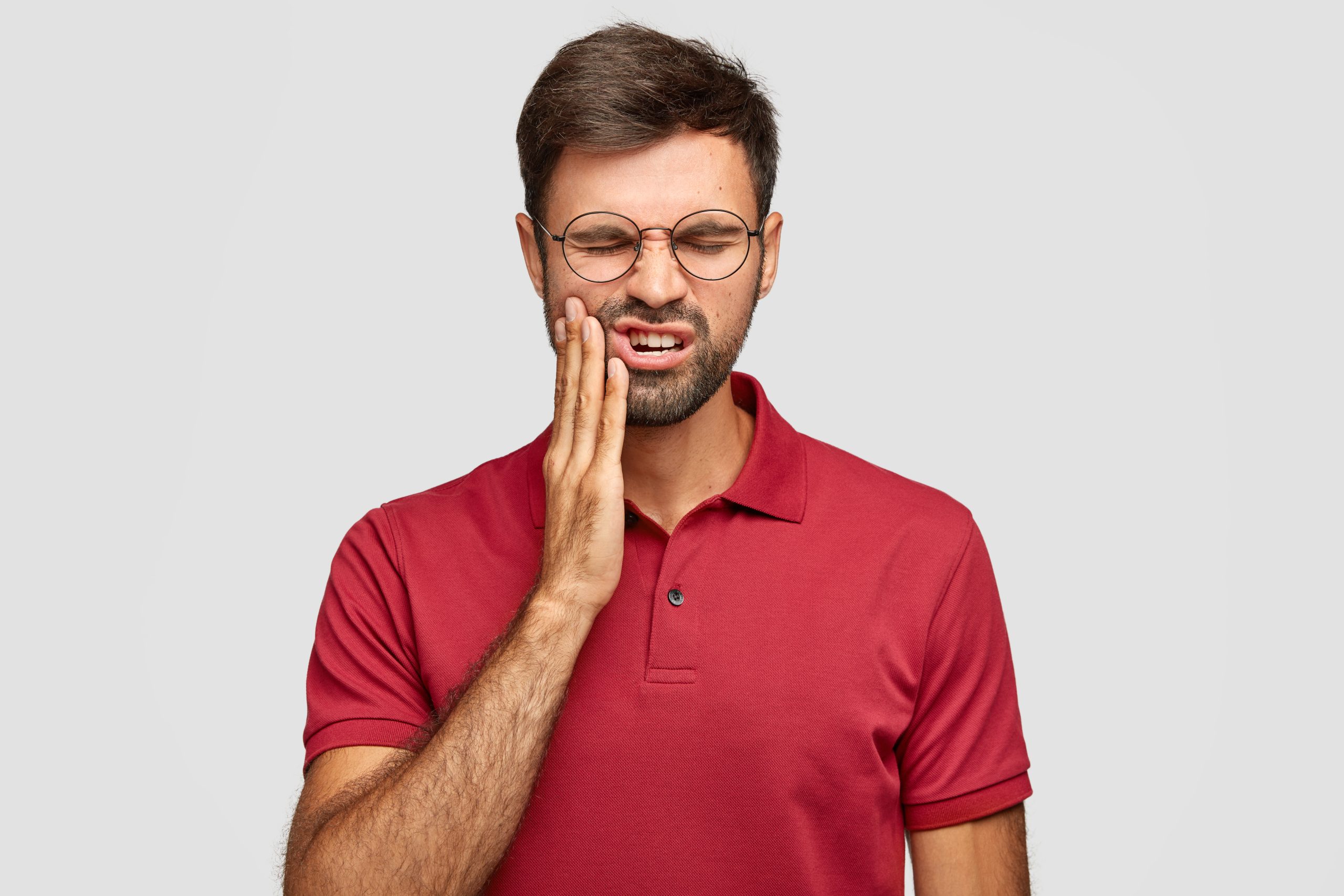 Photo of Caucasian man suffers painful toothache, has rotten tooth, needs to visit dentist, keeps eyes closed from pain, dressed in casual red t shirt, isolated over white background, touches cheek